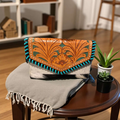 Saddle Bloom Cowhide Crossbody Wallet with hand-tooled floral flap and turquoise whipstitch detail on a wooden table.